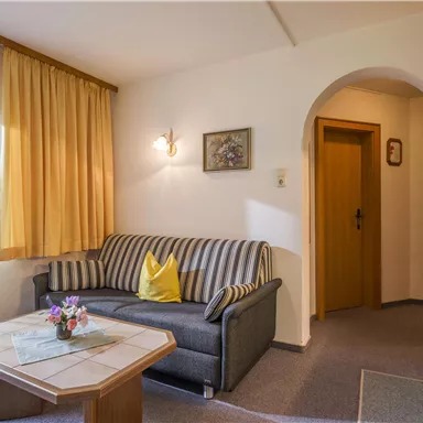 A cozy living room with a gray sofa and a small table. The room has large windows with curtains and a door that leads to another apartment.
