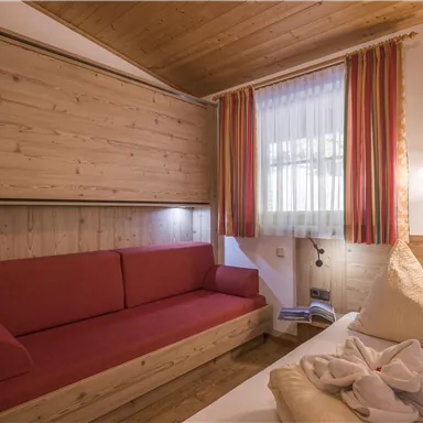 Ein gemütliches Zimmer mit Holzverkleidung, einem roten Sofa und zwei Kopfkissen auf dem Bett. Große Fenster lassen viel Licht herein.