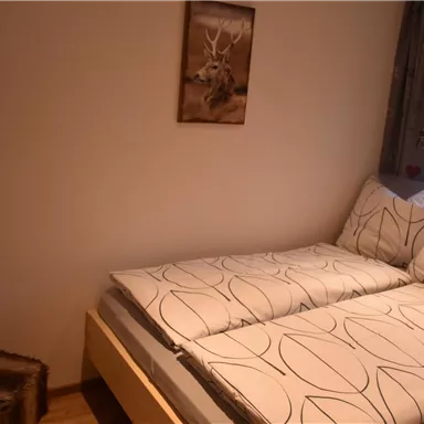 Ein einfaches Schlafzimmer mit einem Bett und einem Stuhl. An der Wand hängt ein Bild eines Rehs.