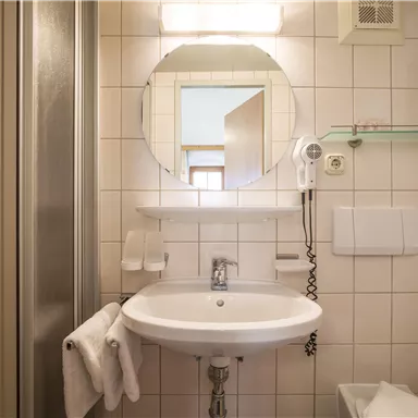 Ein modernes Badezimmer mit einer Dusche, einem Waschbecken und einem großen Spiegel. Die Wände sind gefliest und es gibt einige Handtücher und einen Haartrockner.