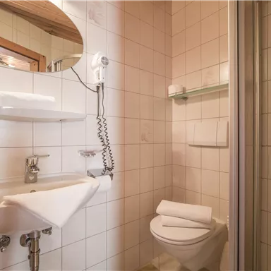 Ein kleines Badezimmer mit weißen Fliesen. Es gibt ein Waschbecken, eine Dusche und eine Toilette.