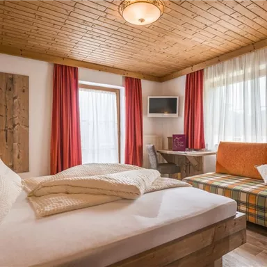 Ein gemütliches Schlafzimmer mit einem Doppelbett und einer Couch. Große Fenster sorgen für viel Tageslicht und eine warme Atmosphäre.