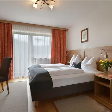 Ein helles Zimmer mit zwei Betten, einem kleinen Tisch und einem Stuhl. Die Fenster sind mit Vorhängen versehen und der Raum ist freundlich dekoriert.