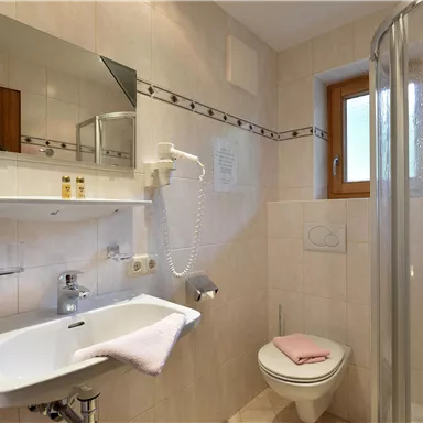 A modern bathroom with a shower, sink, and a toilet. The walls are tiled in light colors, and a window provides daylight.