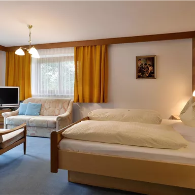 A cozy bedroom with two beds and a television. The room has large windows and warm colors.