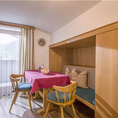 A cozy dining area in a bright kitchen. The table is covered with a red tablecloth and there are chairs made of light wood.