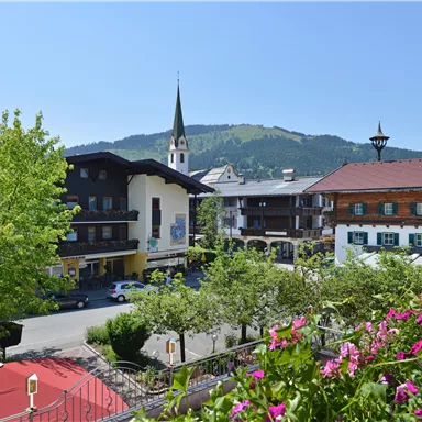 Eine malerische Landschaft mit traditionellen Gebäuden und einem hohen Kirchturm. Im Hintergrund erstrecken sich die Berge unter einem klaren blauen Himmel.
