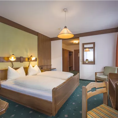 Ein gemütliches Schlafzimmer mit einem Doppelbett, zwei Kissen und Holzdetails. Es gibt einen Tisch, Stühle und ein Fenster mit Vorhängen.