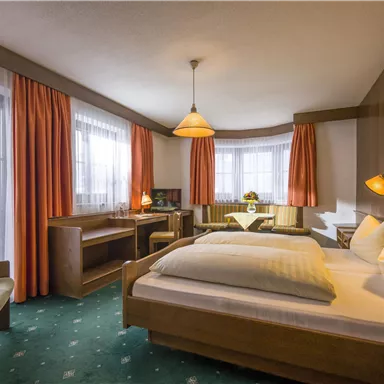 Ein gemütliches Hotelzimmer mit zwei Betten und einem Schreibtisch. Große Fenster mit Vorhängen sorgen für ausreichend Tageslicht.