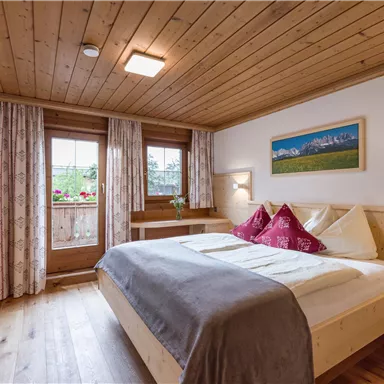 Ein gemütliches Schlafzimmer mit Holzdecke und warmen Farben. Das Zimmer hat ein großes Fenster und dekorative Kissen auf dem Bett.