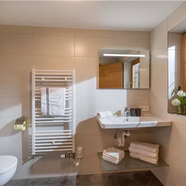 Ein modernes Badezimmer mit einem Waschbecken, einem Spiegel und einem Handtuchhalter. Durch das Fenster kommt natürliches Licht herein, und saubere Handtücher sind ordentlich angeordnet.