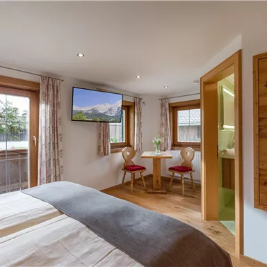 Ein gemütliches Schlafzimmer mit einem großen Bett und einem Fernseher. Es gibt einen kleinen Essbereich mit zwei Stühlen und Fenster, die viel Licht hereinlassen.