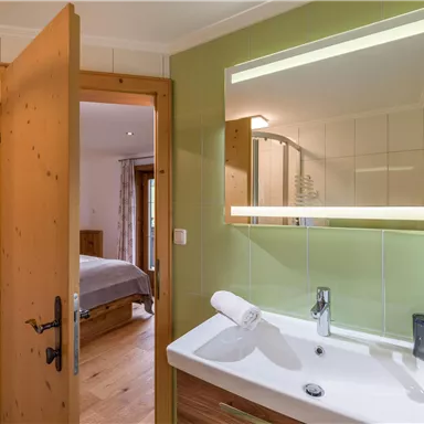 Ein modernes Badezimmer mit einem Waschbecken und großem Spiegel. Im Hintergrund ist ein angegebener Zugang zu einem Schlafzimmer zu sehen.