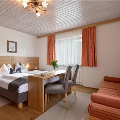 Ein gemütliches Hotelzimmer mit einem Doppelbett, einem Tisch und einer Sitzecke. Die Fenster sind mit Vorhängen in warmen Farben geschmückt.