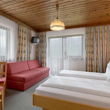 A cozy hotel room with two single beds and a red sofa. The room has large windows and a bright, inviting atmosphere.