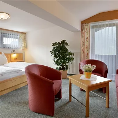 A cozy room with a double bed, red armchairs, and a table. Bright light comes through the windows, and a plant adds to the pleasant atmosphere.
