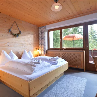 Ein helles Schlafzimmer mit Holzvertäfelung und einem großen Bett. Die Fenster bieten Blick auf einen grünen Außenbereich und einen kleinen Balkon.