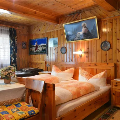 A rustic bedroom with wooden walls and a double bed. On the table, there is a flower vase, and pictures hang on the walls.