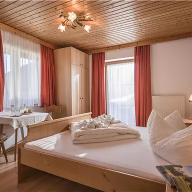 Ein gemütliches Schlafzimmer mit einem Doppelbett und sanften Holzmöbeln. Große Fenster sorgen für viel Tageslicht und eine angenehme Atmosphäre.