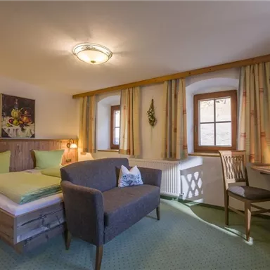 Ein gemütliches Hotelzimmer mit einem Doppelbett, einem Sessel und einem kleinen Schreibtisch. Die Wände sind in warmen Farben gehalten, und große Fenster lassen viel Tageslicht herein.