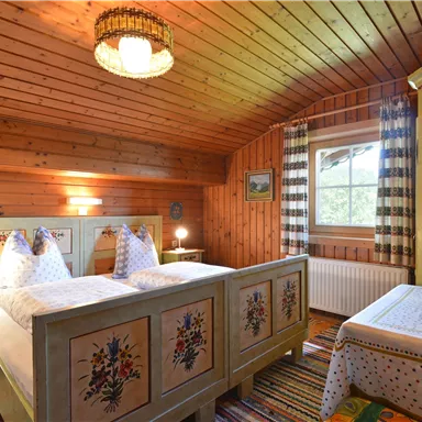 A cozy bedroom with a wooden ceiling and rustic furnishings. Two beds with decorative headboards and a sunny window create an inviting atmosphere.