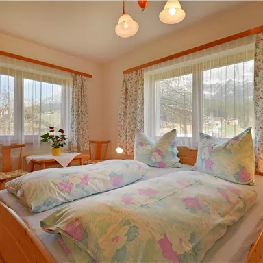 Ein gemütliches Schlafzimmer mit einem Doppelbett und bunten Kissen. Große Fenster lassen viel Licht herein und der Raum ist hell und freundlich eingerichtet.