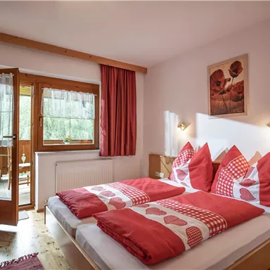 Ein gemütliches Schlafzimmer mit zwei Betten und roten Kissen. Große Fenster öffnen sich zu einem Balkon mit Blick ins Freie.