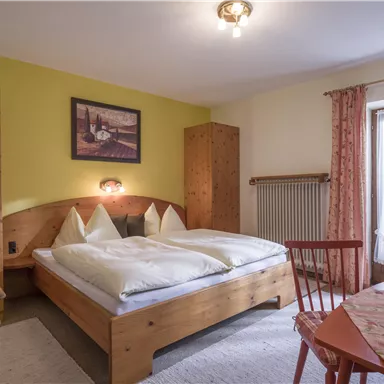 Ein gemütliches Schlafzimmer mit einem Doppelbett und Holzmöbeln. Große Fenster lassen viel Tageslicht hereinfällen und bieten einen Blick nach draußen.