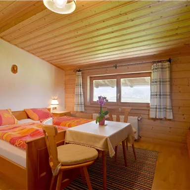 A cozy room with a wooden ceiling, two beds, and a table. Curtains are at the window, and a small bouquet of flowers decorates the table.