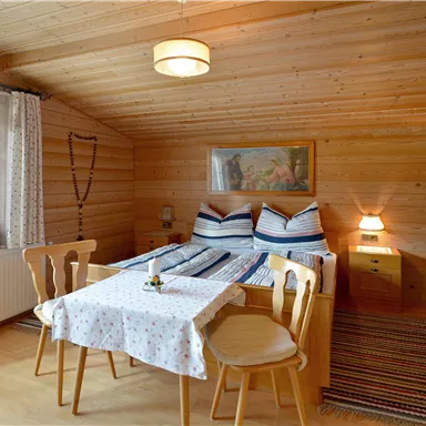 A cozy room with wooden walls, a double bed, and a small dining area. A table with a blue tablecloth and two chairs is in the center of the room.