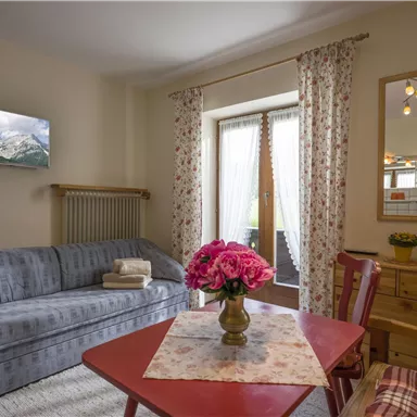 Ein gemütliches Wohnzimmer mit einem grauen Sofa, einem Tisch und einem Blumenstrauß. Große Fenster sorgen für viel Tageslicht und eine einladende Atmosphäre.