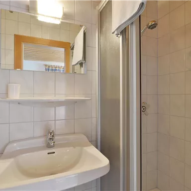 Ein modernes Badezimmer mit einer Dusche, einem Waschbecken und einem Spiegel. Die Wände sind mit hellen Fliesen gefliest.