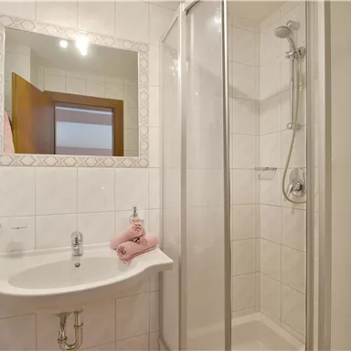 A modern bathroom with a shower and a sink. The walls are tiled in light colors, and there are pink towels on the sink.