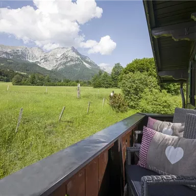 Ein wunderschöner Ausblick von einem Balkon auf grüne Wiesen und beeindruckende Berge. Die gemütlichen Sessel laden zum Entspannen ein.