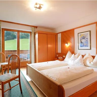 A cozy hotel room with two beds, wood paneling, and a view of the green landscape. The room is bright and cheerfully furnished, with a table and chairs.
