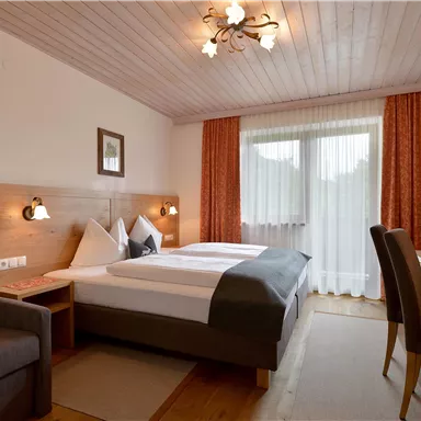 Ein gemütliches Hotelzimmer mit einem Doppelbett, einer Sitzecke und einem Schreibtisch. Große Fenster sorgen für viel Tageslicht und eine helle Atmosphäre.
