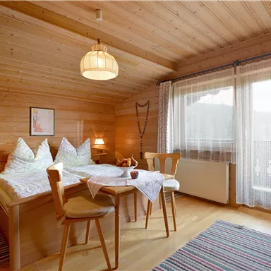A cozy room with wooden walls and two beds. There is a table with chairs and large windows that let in a lot of light.
