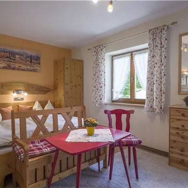 Ein gemütliches Zimmer mit einem Doppelbett und einem kleinen Esstisch. Helle Vorhänge und ein großes Fenster sorgen für eine freundliche Atmosphäre.