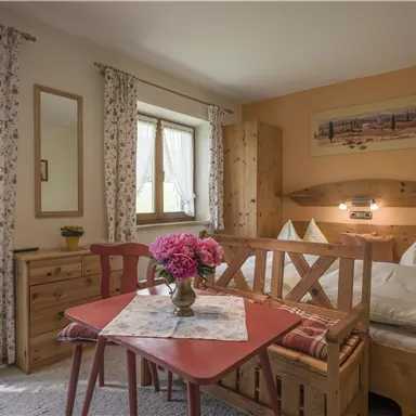 Ein gemütlicher Raum mit einem Doppelbett und einem kleinen Tisch. Die Fenster sind mit hellen Vorhängen geschmückt und frische Blumen stehen in einer Vase.