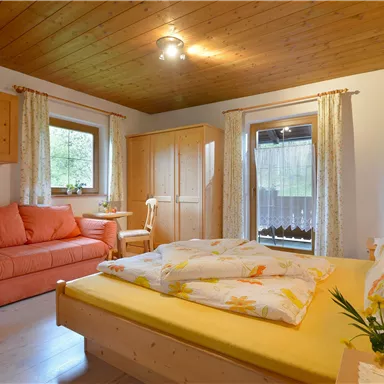 Ein gemütliches Schlafzimmer mit einem Doppelbett, einem Sofa und Holzregalen. Große Fenster lassen viel Licht herein und die Einrichtung ist freundlich und einladend.