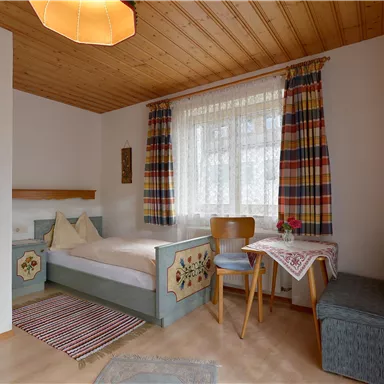 Ein gemütliches Zimmer mit Holzdecke und bunten Vorhängen. Es enthält ein Einzelbett, einen Tisch und einen Sessel.