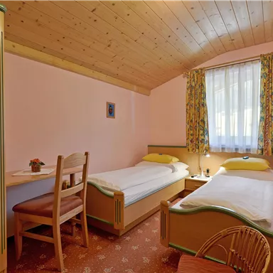 Ein gemütliches Zimmer mit zwei Einzelbetten und einem kleinen Tisch. Die Wände sind rosa und die Fenster haben bunte Gardinen.