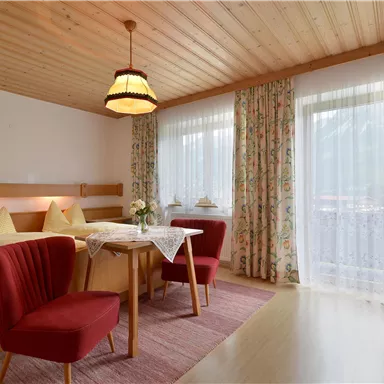 Ein gemütliches Zimmer mit einem Doppelbett und rotem Sitzmöbeln. Große Fenster sorgen für viel Tageslicht und einen schönen Ausblick.