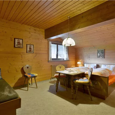 A cozy room with wooden walls and a wooden floor. It contains a double bed, a table, and a sitting area.