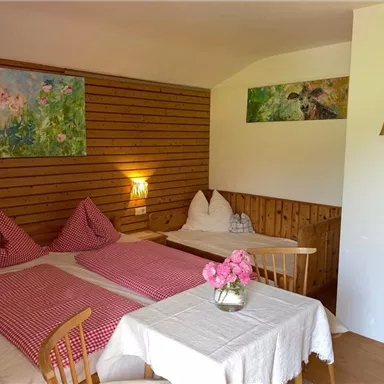 Ein gemütliches Zimmer mit Holzverkleidung und zwei Betten. Auf dem Tisch steht eine Vase mit Rosen und es hängen bunte Bilder an der Wand.