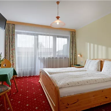 A cozy bedroom with two beds and a table. Large windows provide plenty of daylight.