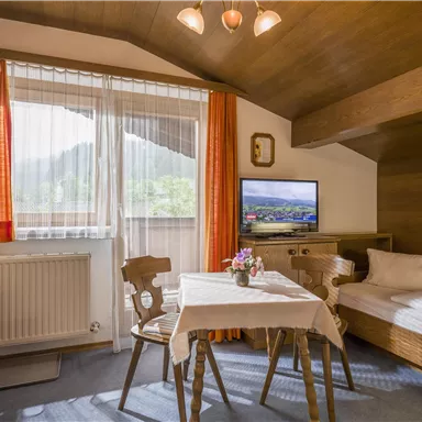 A cozy room with wood paneling and a large window. There is a table, chairs, and a television, as well as a single bed.