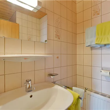 A bathroom with white tiles, a functional sink, and a wall mirror. Towels in yellow and blue hang next to it.