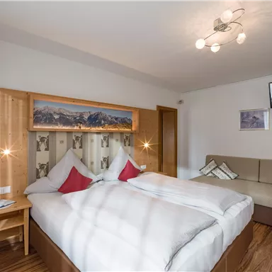 A cozy room with a double bed and two red pillows. On the wall, there are pictures of mountains and a television mounted.