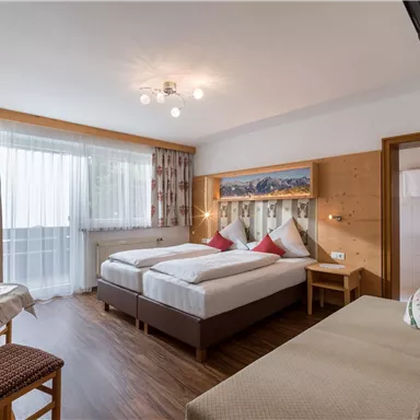 A cozy bedroom with two single beds, a couch, and a table. Large windows provide plenty of daylight and a view outside.